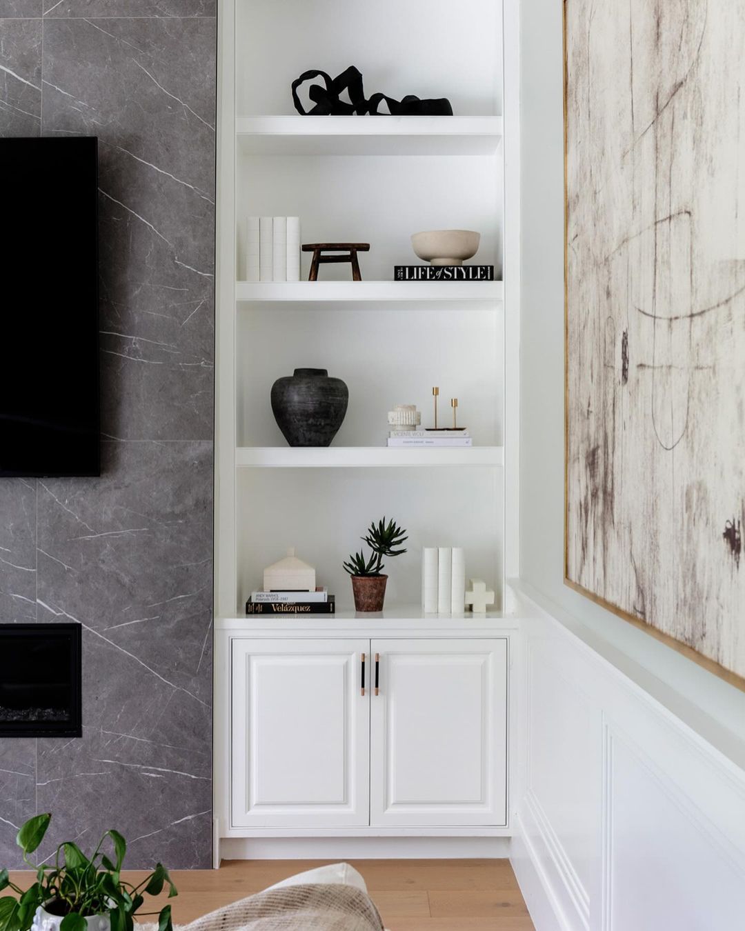 White bookcase with decorative objects, books, and a plant. Modern interior design with a fireplace and abstract art on the wall.