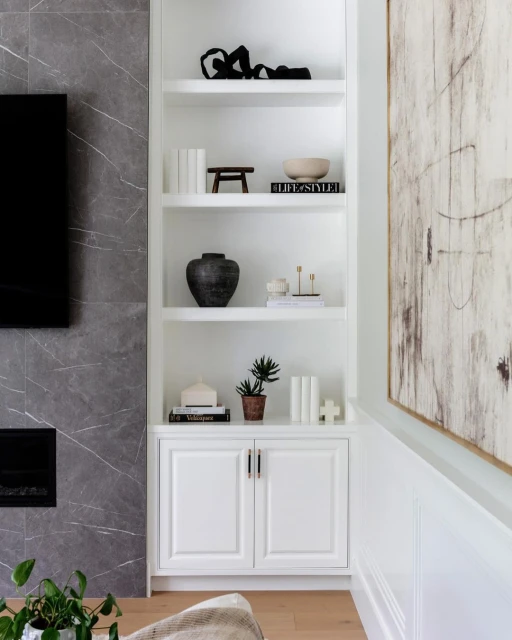 White bookcase with decorative objects, books, and a plant. Modern interior design with a fireplace and abstract art on the wall.