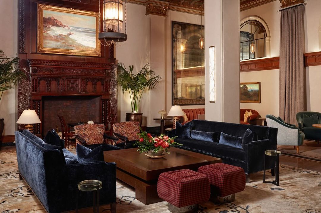 Ornate hotel lobby interior with blue sofas, wood tables, and a vintage fireplace. Artwork and plants enhance the luxurious setting.