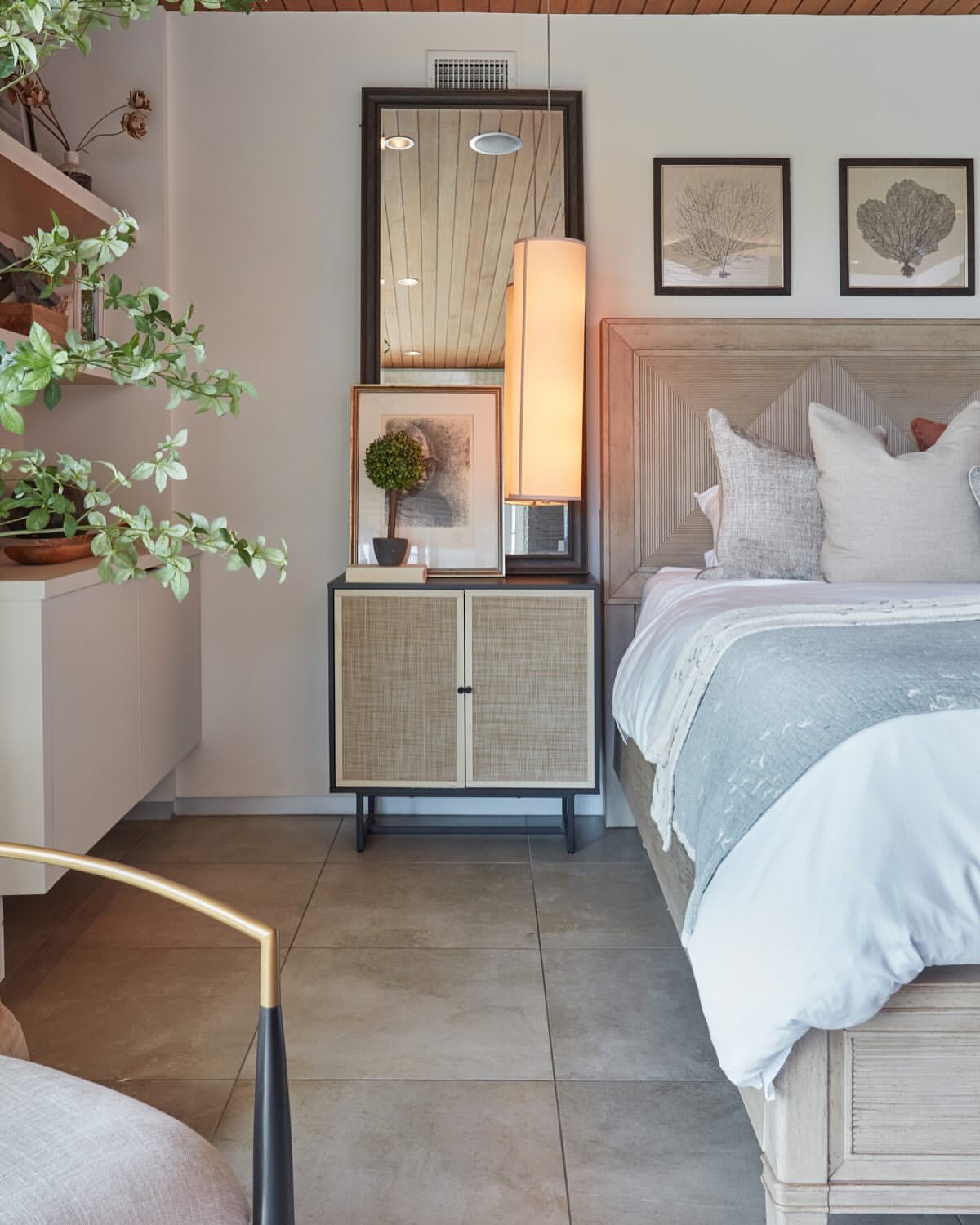 Bedroom interior with a bed, bedside table with a mirror, lamps, artwork, and decorative plants. Light neutral color palette.