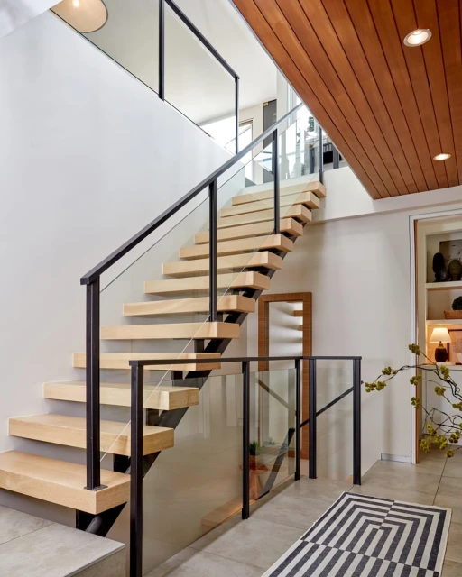 Modern floating staircase with light wood steps, black metal railings, and glass panels in a brightly lit home interior.