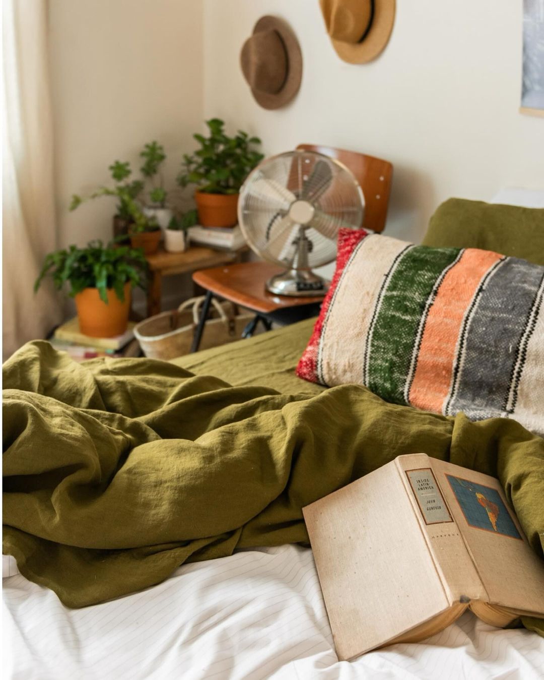 Bedroom interior with a green linen-covered bed, an open book, a patterned pillow, plants, and vintage decor create a cozy, inviting scene.