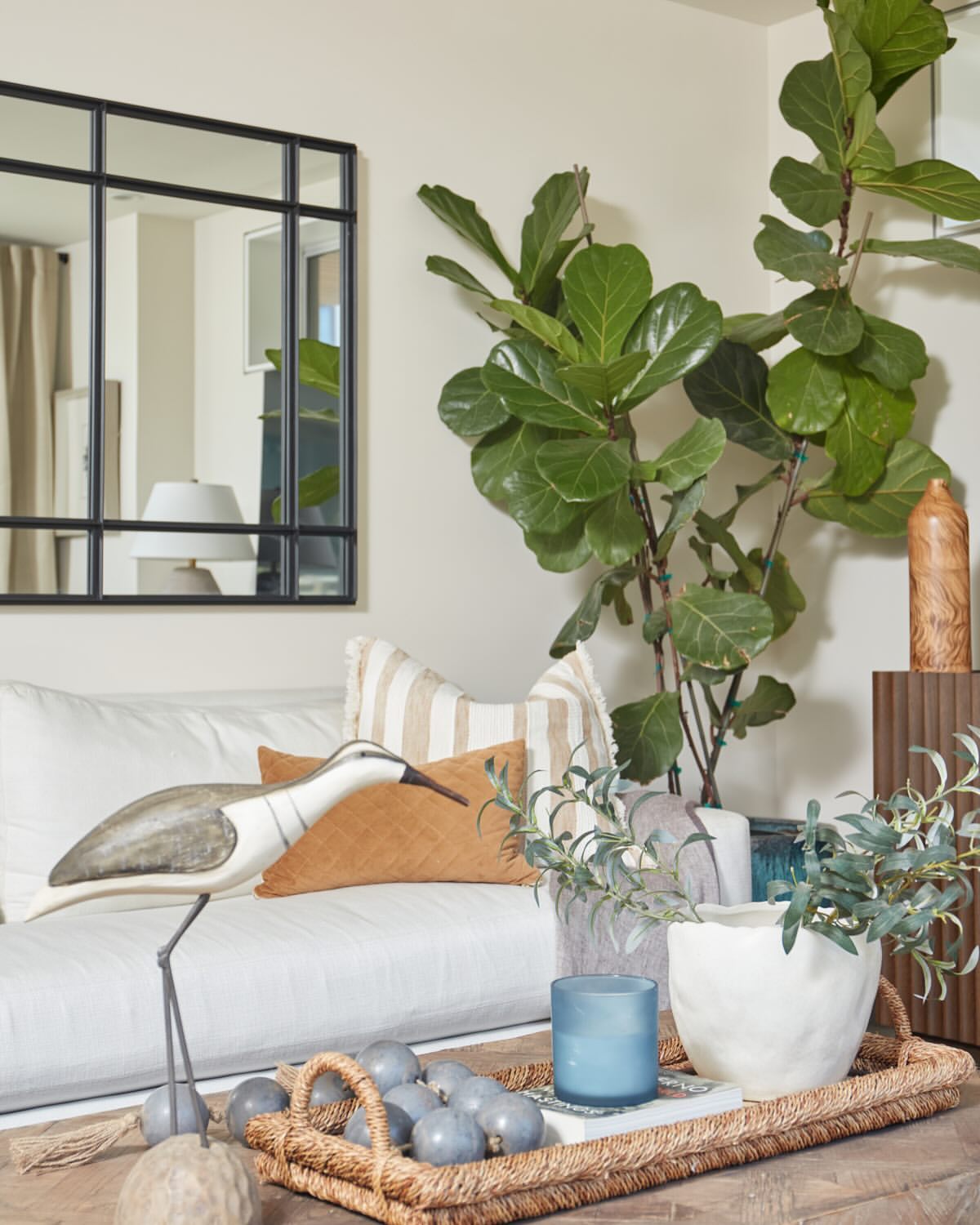 Interior of a living room featuring a decorative bird sculpture, sofa with pillows, plants, and a wall mirror in a neutral color scheme.