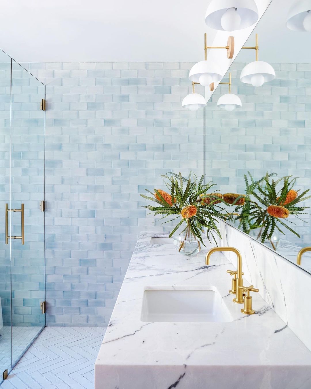 Contemporary bathroom interior with white marble countertop, gold fixtures, glass shower, blue-tiled walls, and decorative floral arrangement.