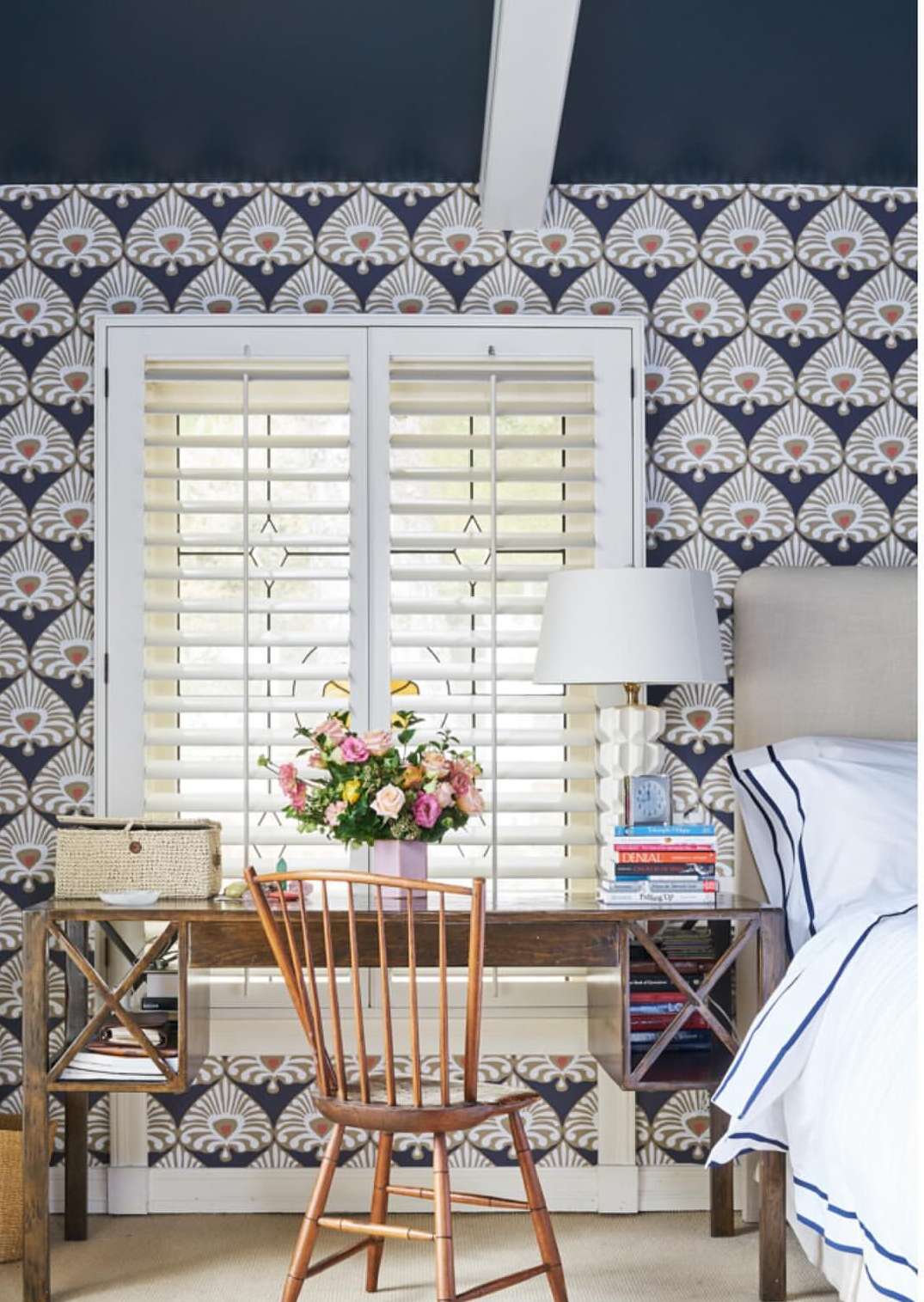 Bedroom interior featuring a wooden desk adorned with a floral arrangement, books, and a lamp, beside a bed with striped bedding.