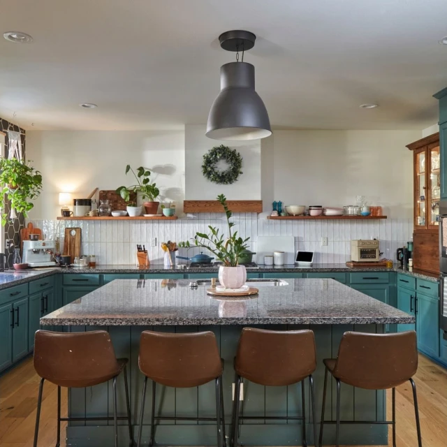 Interior shot of a kitchen with teal cabinetry, a large island with brown stools, plants, and a black pendant light overhead.