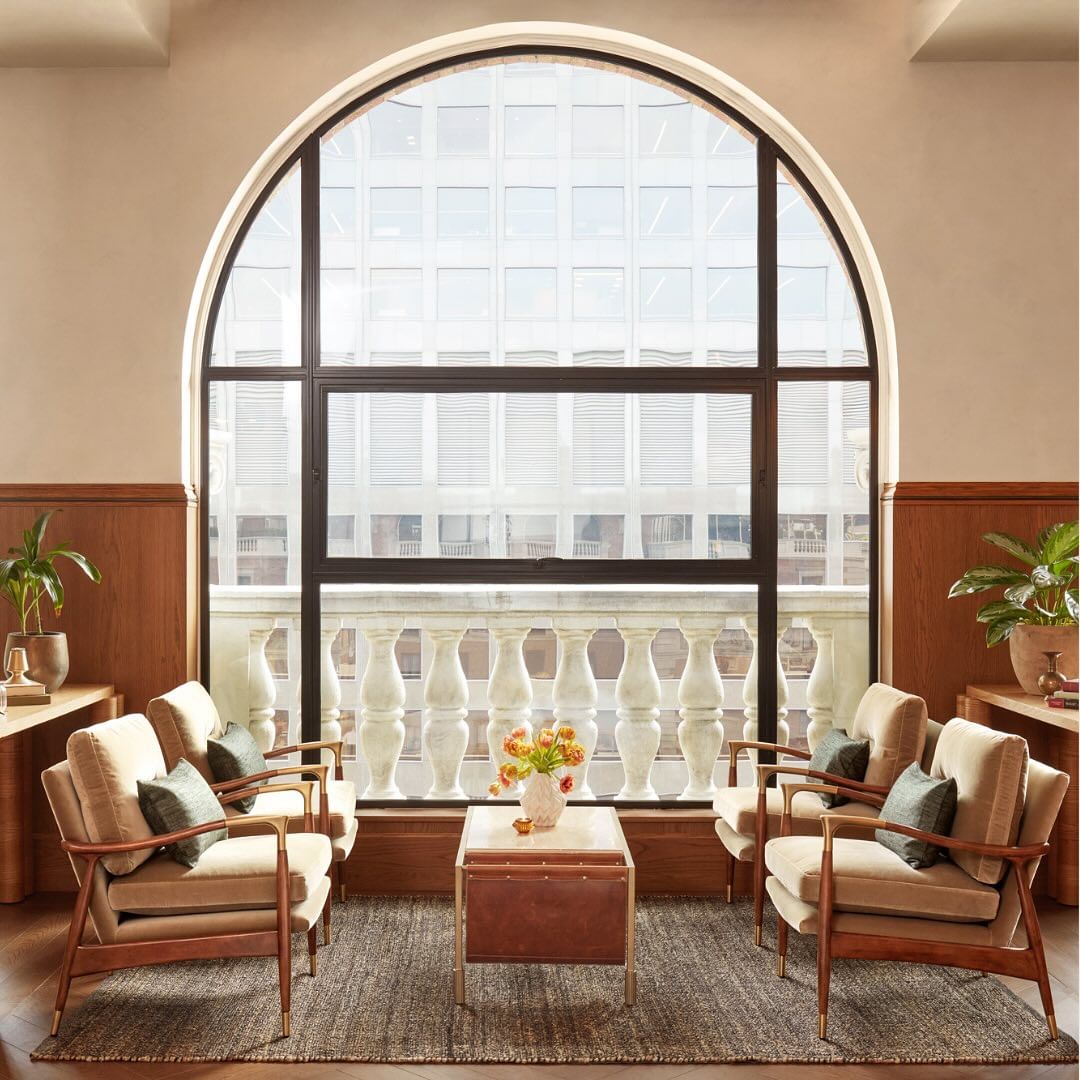 Interior view of a modern lounge area with four beige armchairs, a central coffee table with flowers, and a large arched window providing natural light.