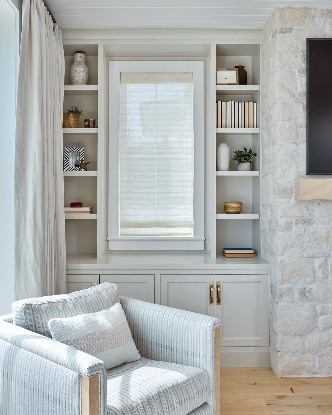 Interior view of a neutral-toned living room featuring a striped armchair, custom built-in shelving, decor, and a stone fireplace.