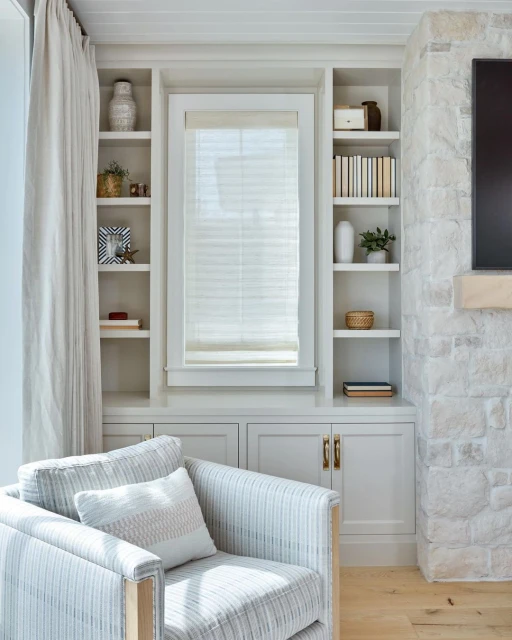 Interior view of a neutral-toned living room featuring a striped armchair, custom built-in shelving, decor, and a stone fireplace.