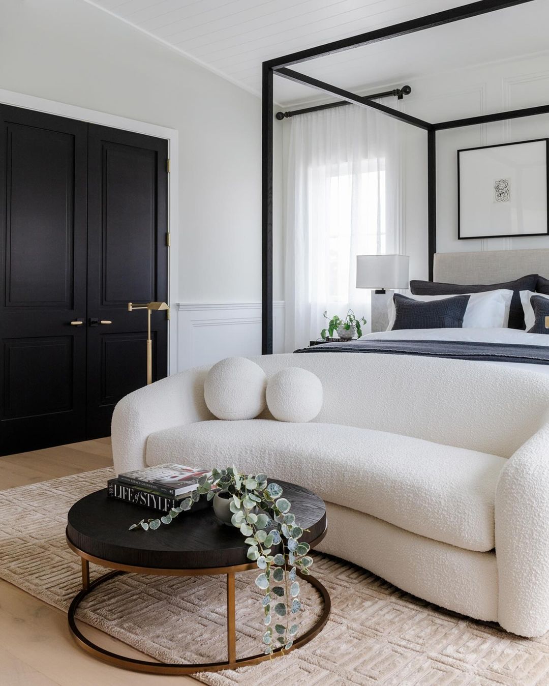 Interior of bedroom featuring a white sofa, a round coffee table with books and greenery, and a black canopy bed in a neutral palette.