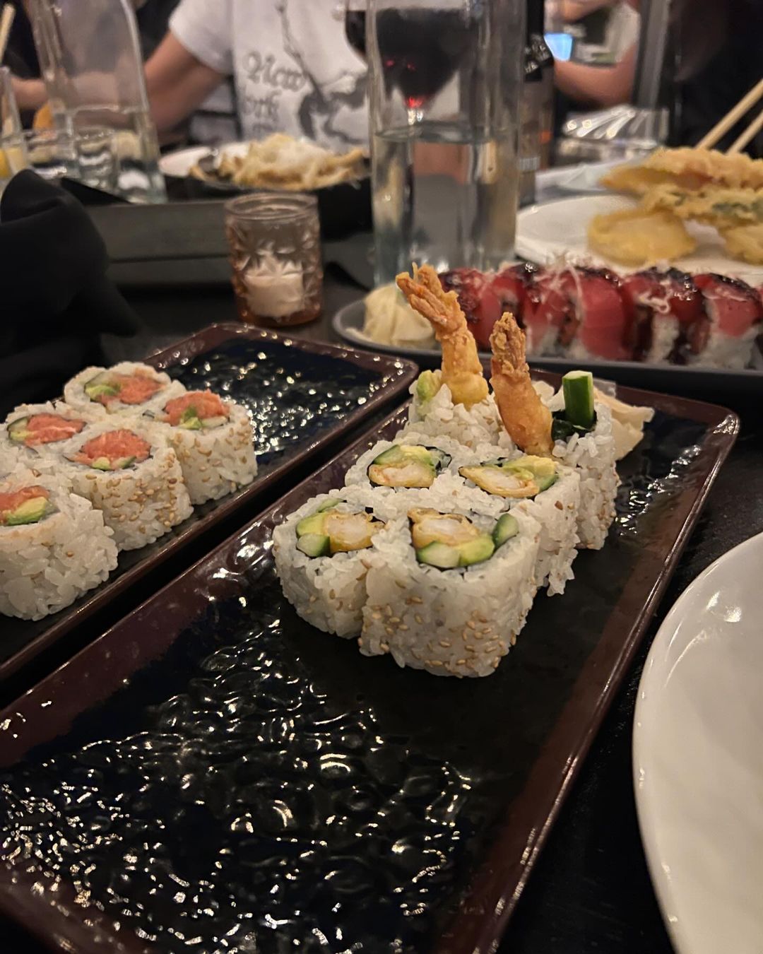 A dark table setting displays two rectangular plates of sushi rolls, with shrimp tempura and avocado accents, plus side dishes and drinks.