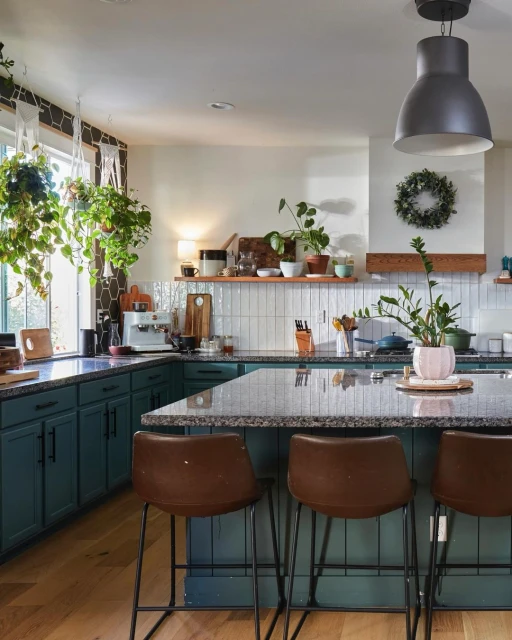Indoor kitchen scene with teal cabinets, granite countertops, brown leather stools, and hanging potted plants near a window.