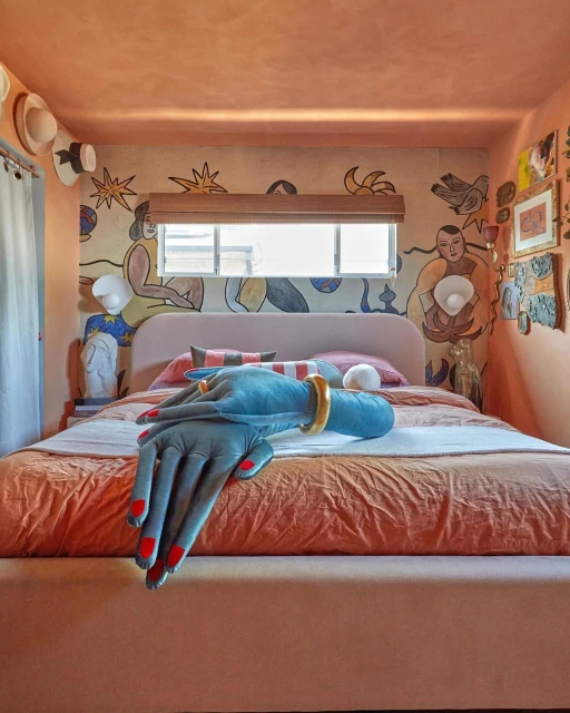 Whimsical bedroom interior featuring an art-covered wall, window with light filtering through, and a decorative hand sculpture on the bed.