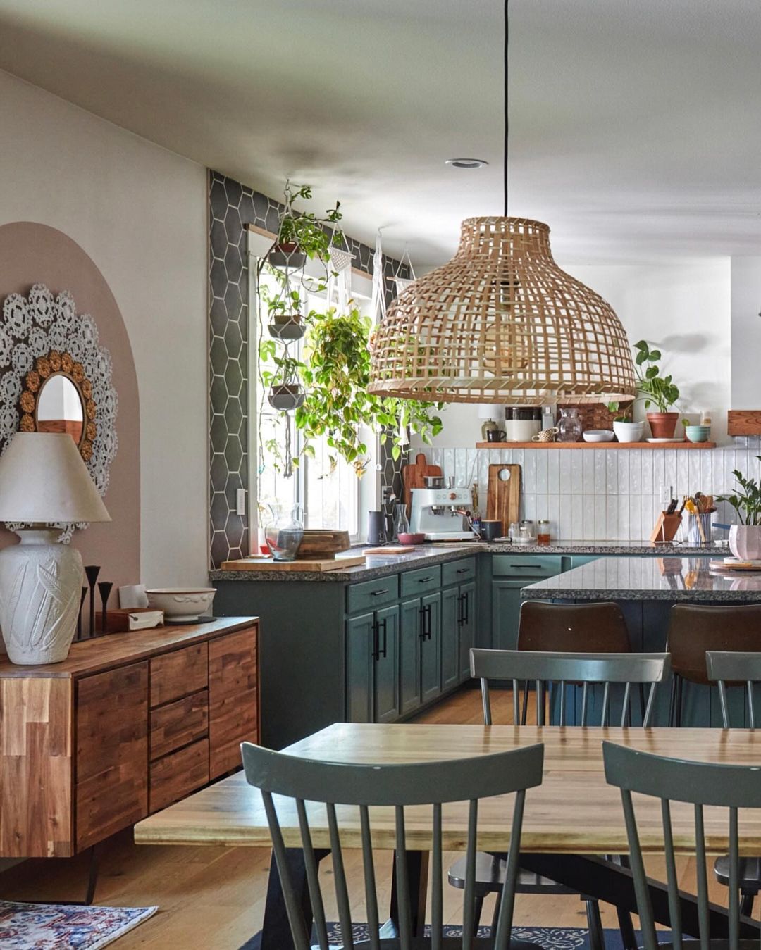 A bright kitchen features green cabinets, a wooden dining table, an island with bar stools, hanging plants, and a rattan pendant light.