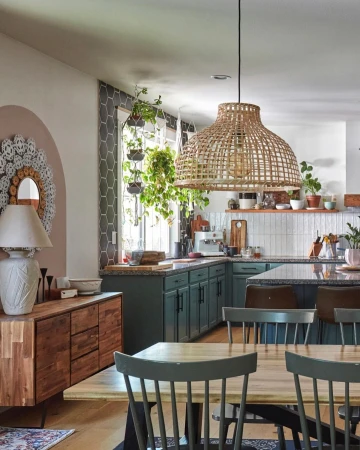 A bright kitchen features green cabinets, a wooden dining table, an island with bar stools, hanging plants, and a rattan pendant light.