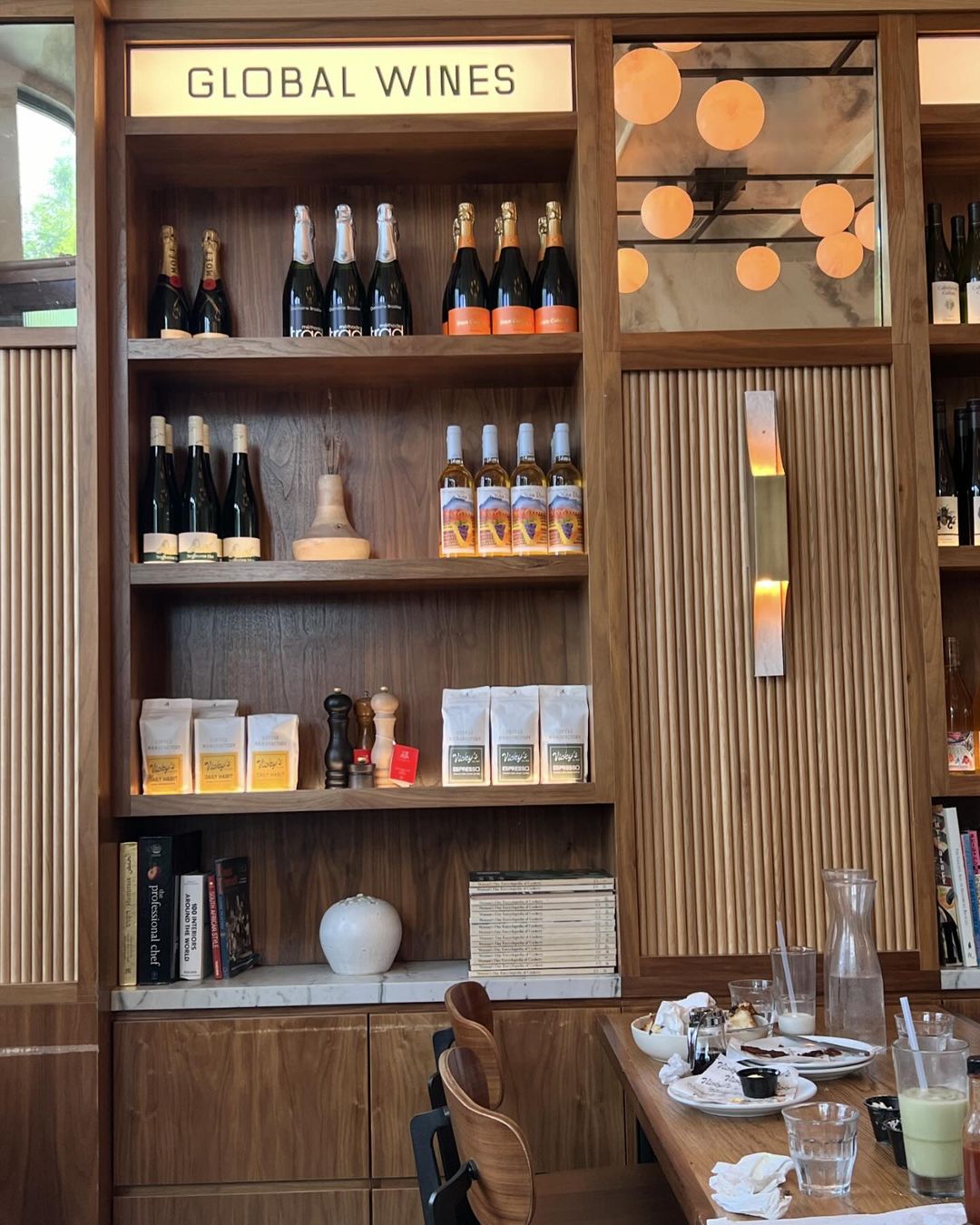 Interior shot of a wooden shelf with a variety of wine bottles, coffee bags, and books, lit by overhead lights and a decorative wall sconce.