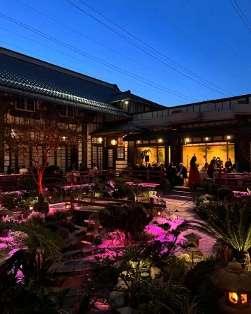 Outdoor garden party at night with guests seated at tables, illuminated by lanterns and string lights under a dark blue sky.