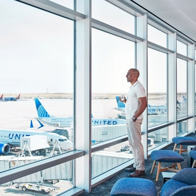 Adult male standing near an airport window, looking out at the planes, holding a coffee mug, wearing a white polo and light-colored pants.