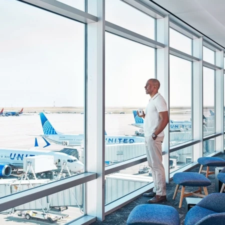 Adult male standing near an airport window, looking out at the planes, holding a coffee mug, wearing a white polo and light-colored pants.