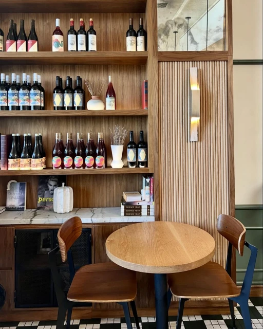 Interior shot of a restaurant or bar with a wooden shelf stocked with wine bottles, above a round table with chairs and a checkered floor.