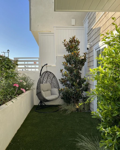 Outdoor patio featuring a hanging egg chair, lush green artificial turf, a small magnolia tree, and a white privacy wall in daylight.