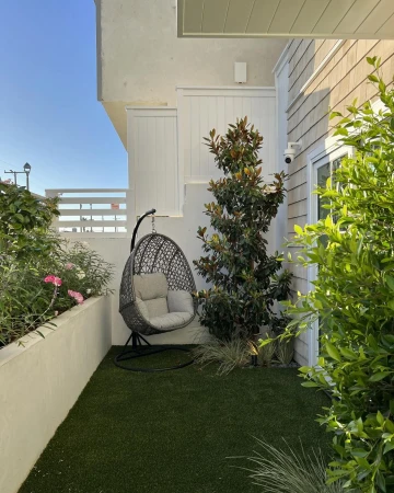 Outdoor patio featuring a hanging egg chair, lush green artificial turf, a small magnolia tree, and a white privacy wall in daylight.