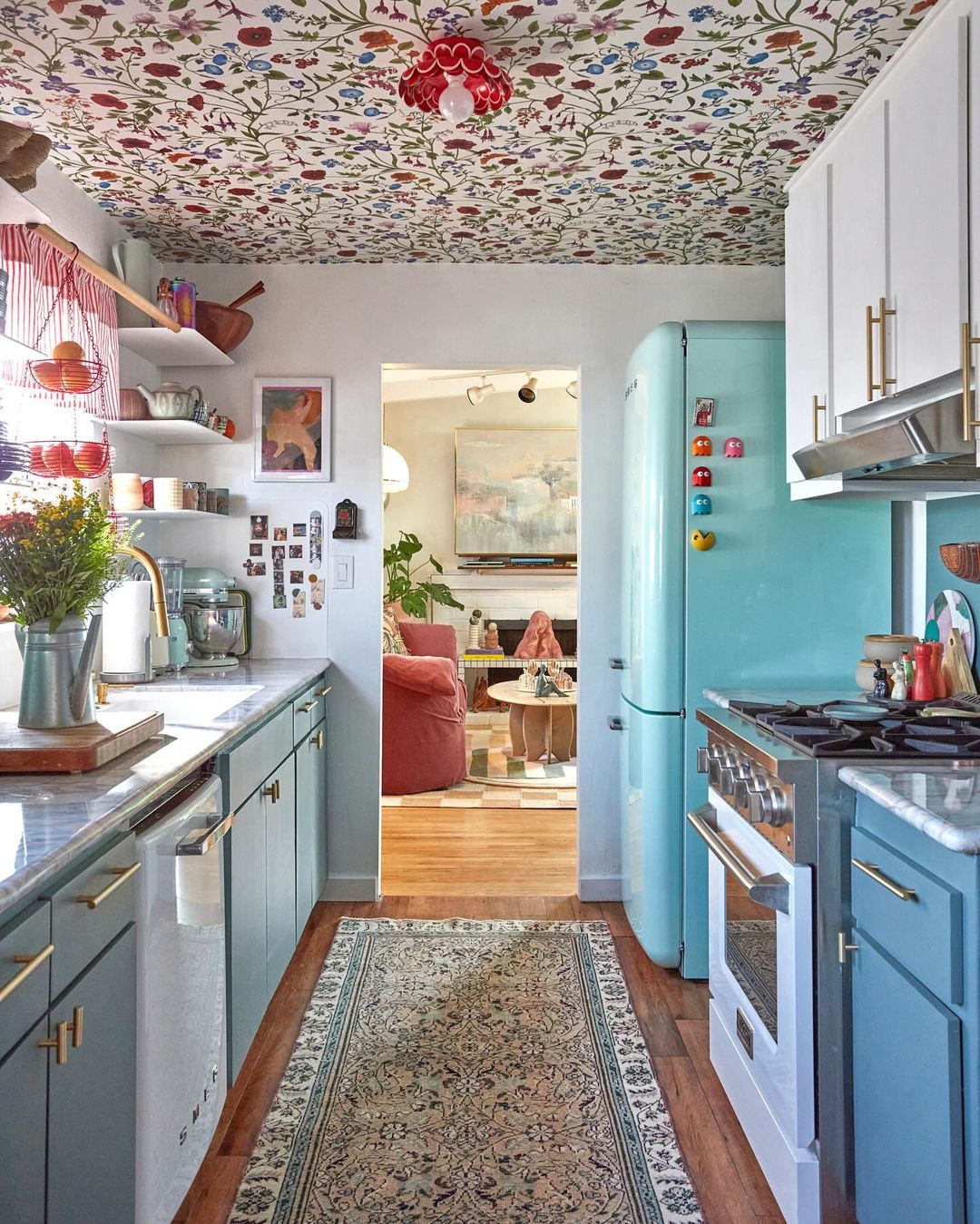 A charming kitchen view shows pastel-colored cabinets, floral wallpaper on the ceiling, and an open doorway to a living area.