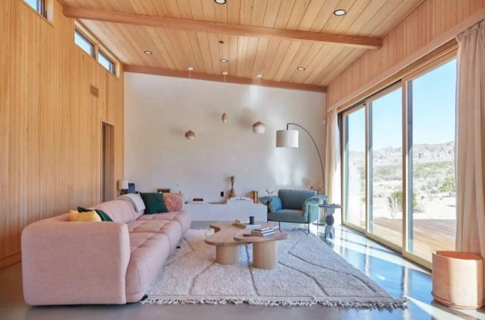 Living room interior featuring a pink couch, wooden furniture, white walls, and a large window with an outdoor view of a desert landscape.