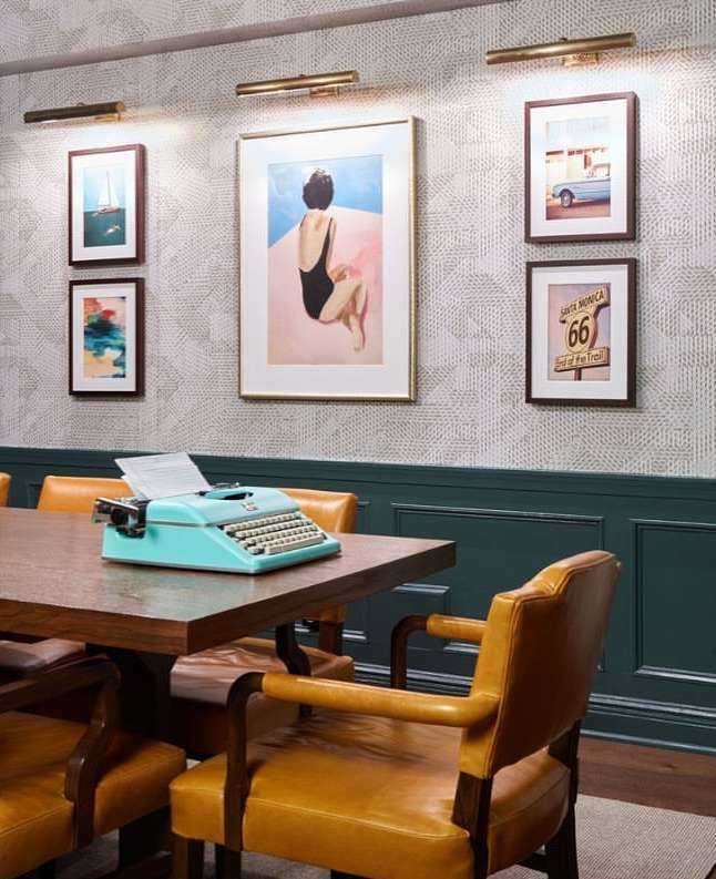 Indoor dining area with a wooden table, mustard-colored chairs, and a blue typewriter. Artwork and wall lighting complete the decor.
