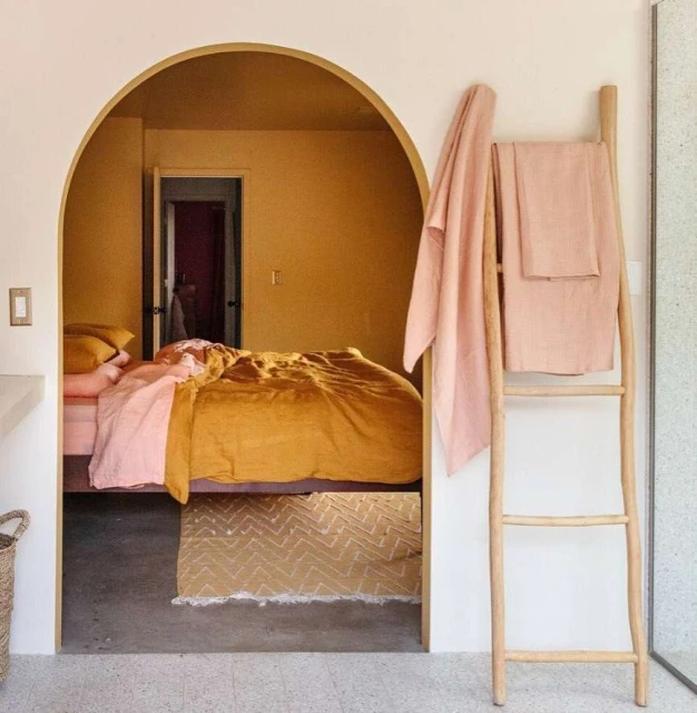 Bedroom scene with mustard-colored bedding, a pink blanket, and a wooden ladder with pink towels in a brightly lit interior space.