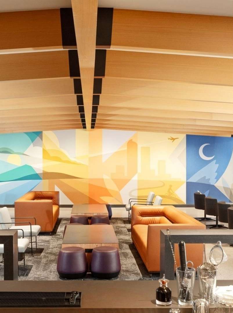 Interior of an airport lounge area with comfortable seating, multiple tables, a mural on the wall, and a bar with glassware.