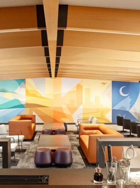 Interior of an airport lounge area with comfortable seating, multiple tables, a mural on the wall, and a bar with glassware.
