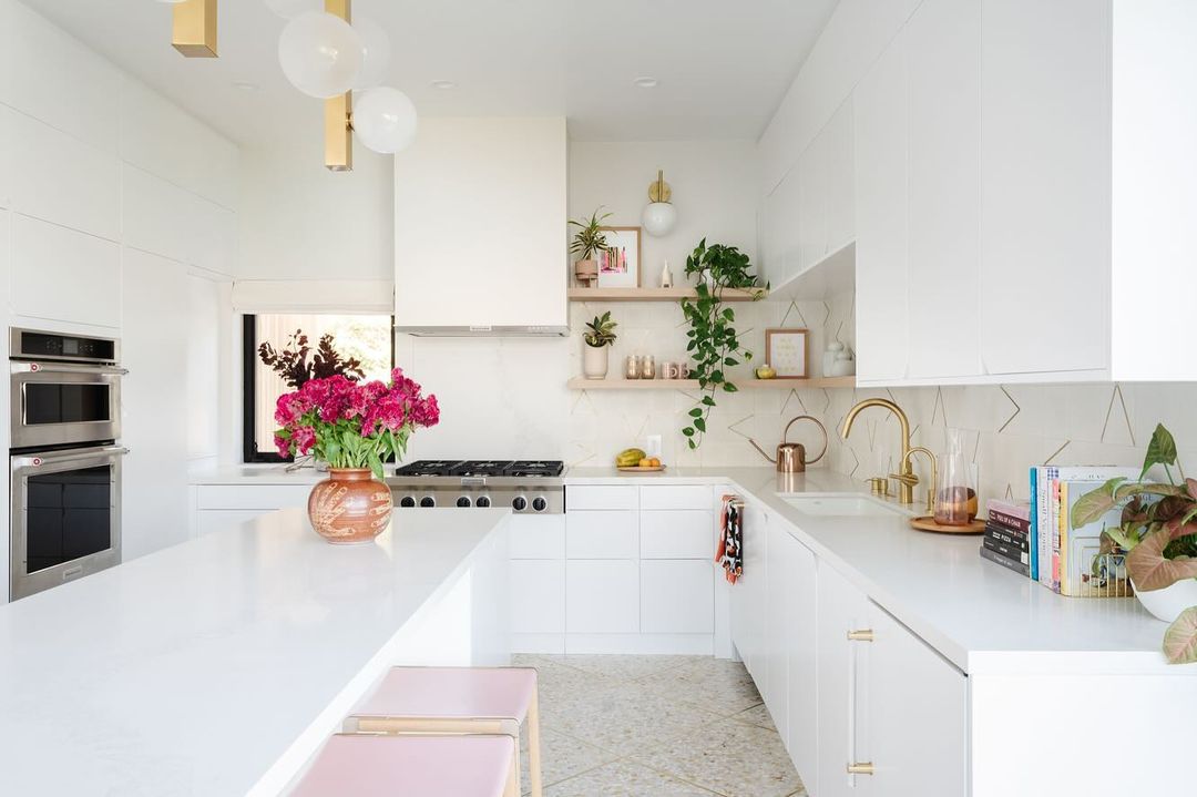 Bright, modern kitchen interior with white cabinets, countertops, stainless steel appliances, pink stools, and fresh flowers in a vase.