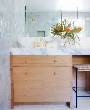 Bathroom interior showcasing a light wood vanity with a marble countertop, decorative floral arrangement, gold fixtures, and a woven stool.