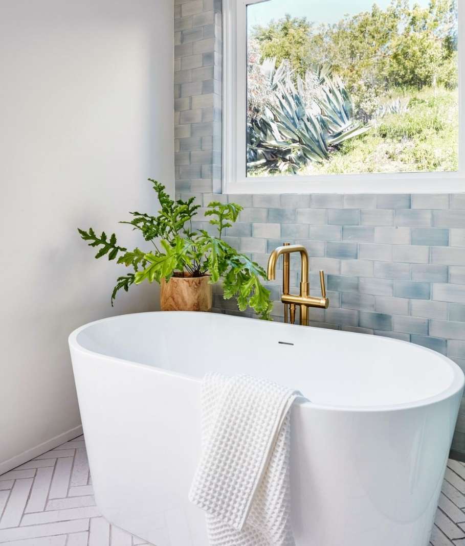 Bright bathroom featuring a white oval tub, gold faucet, houseplant, and a window overlooking a green hillside landscape.