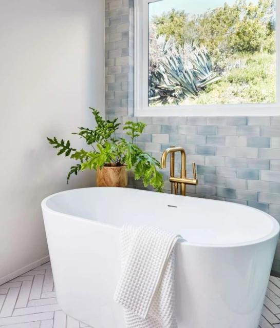 Bright bathroom featuring a white oval tub, gold faucet, houseplant, and a window overlooking a green hillside landscape.