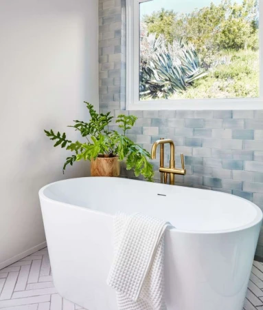 Bright bathroom featuring a white oval tub, gold faucet, houseplant, and a window overlooking a green hillside landscape.