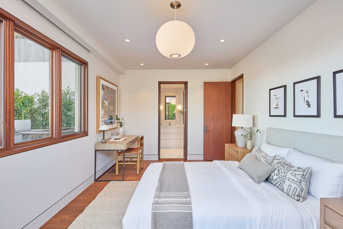 Bright and airy bedroom interior with a white bed, wood accents, a desk, and natural light from a window. Modern home design.