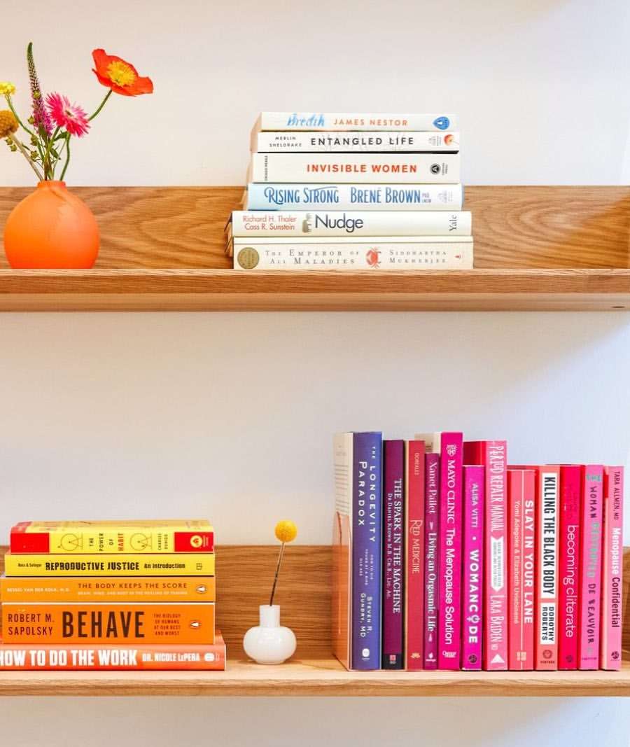 Two wooden shelves with stacks of colorful books, a vase of flowers, and a decorative white vase against a neutral wall.