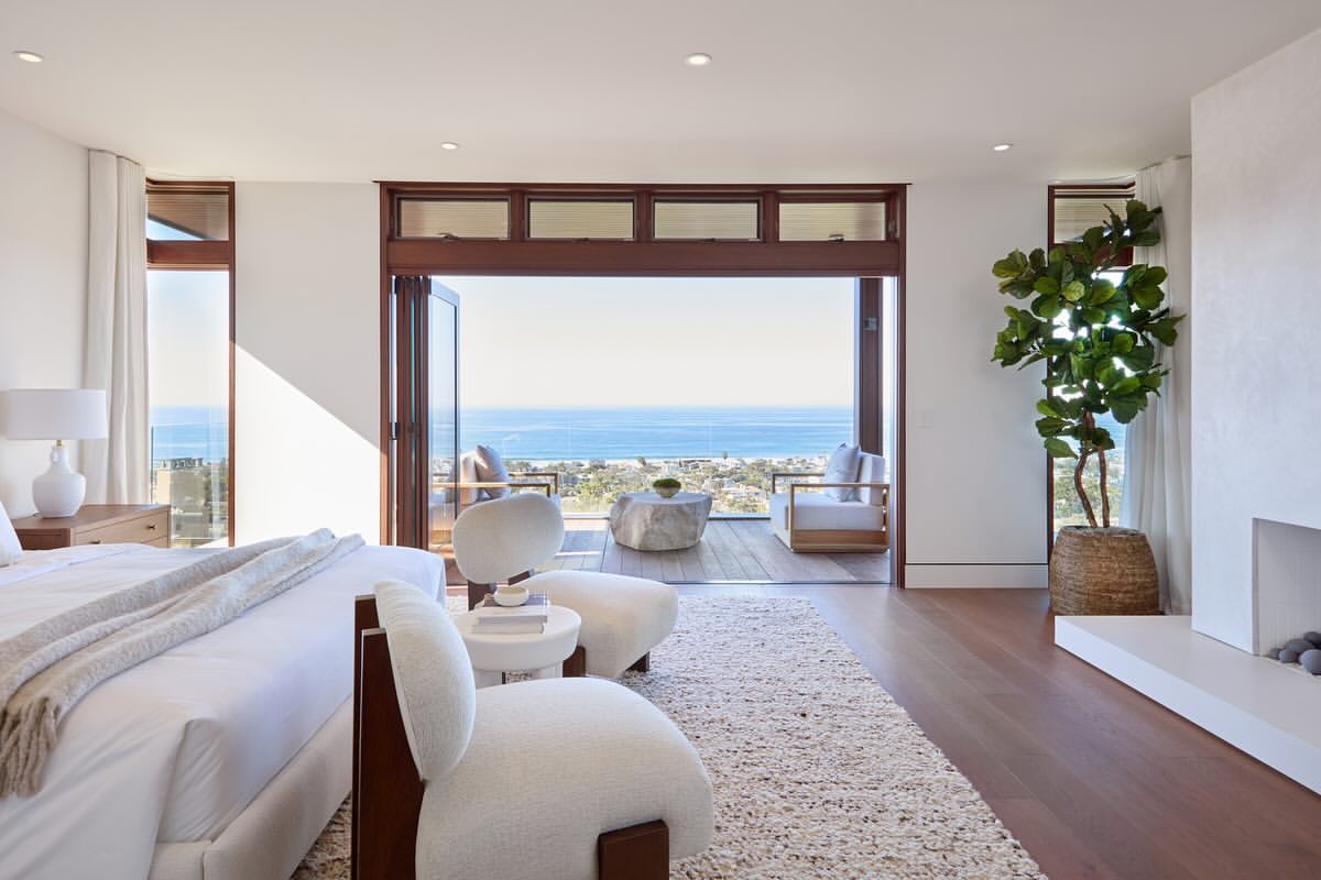 Bright bedroom with neutral tones featuring a king bed, armchairs, fireplace, and an ocean view through the folding glass doors.