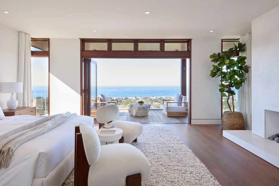 Bright bedroom with neutral tones featuring a king bed, armchairs, fireplace, and an ocean view through the folding glass doors.