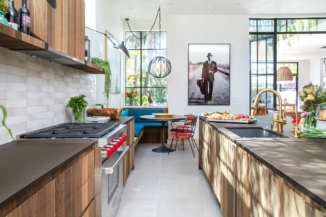 Warm modern kitchen interior with wood cabinetry, dark countertops, and a dining nook beside a window and a print.