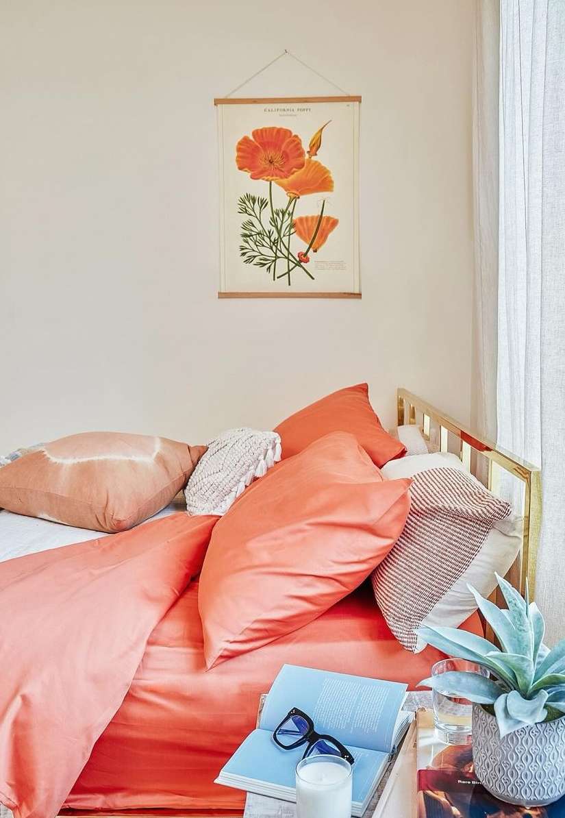 Cozy indoor scene featuring an orange flower botanical print, pillows, and a book with glasses atop a bed in a brightly lit room.