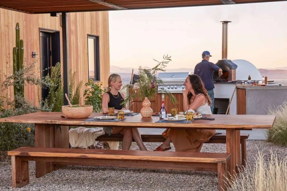 Two women at a wooden table outside a modern house, with a man near a pizza oven, featuring a rustic picnic table and outdoor kitchen.