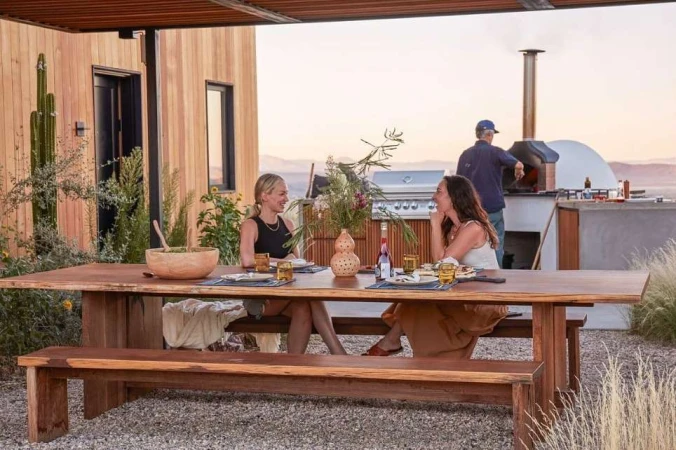 Two women at a wooden table outside a modern house, with a man near a pizza oven, featuring a rustic picnic table and outdoor kitchen.