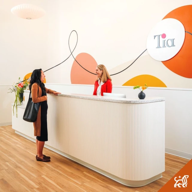 Two women converse at a modern, minimalist reception desk, with a large logo and abstract wall art in a bright, inviting healthcare setting.