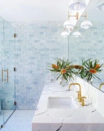 Modern bathroom interior featuring a white marble vanity with a gold faucet, pale blue tiled walls, and brass wall sconces.