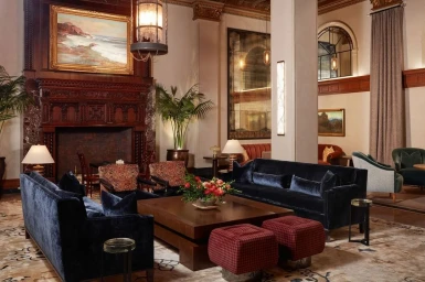 Luxurious hotel lobby interior featuring dark velvet sofas, patterned armchairs, and a large wooden fireplace with a seascape painting above.