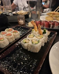 Close up of sushi rolls on dark rectangular plates on a restaurant table with drinks and plates of food in the background