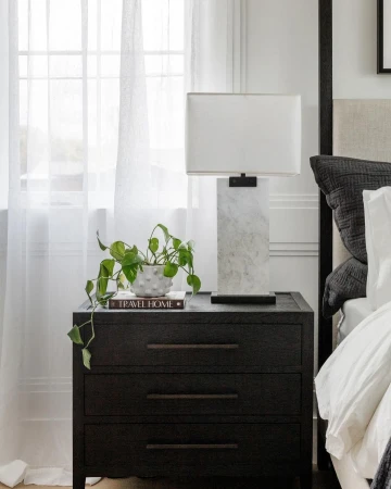 Dark wooden nightstand featuring a marble lamp and a potted trailing plant in a bedroom with sheer curtains.